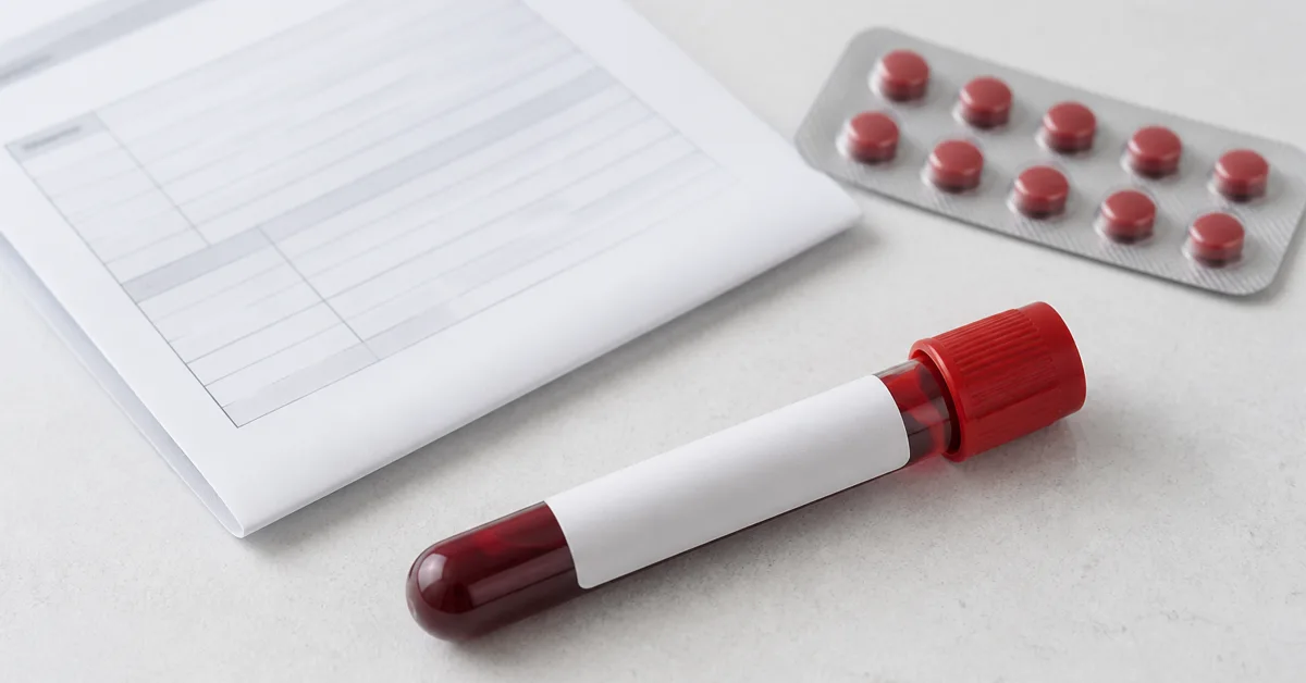 Blutentnahmeröhrchen mit Etikett neben einem Laborformular und einer Vitamin-B12-Packung auf hellem Untergrund